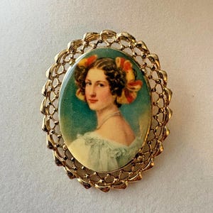 Op de afbeelding: Ovale vintage broche met een portret van een vrouw. Het portret toont een vrouw met krullend haar versierd met oranje en gele linten. De broche heeft een goudkleurig filigraan frame.