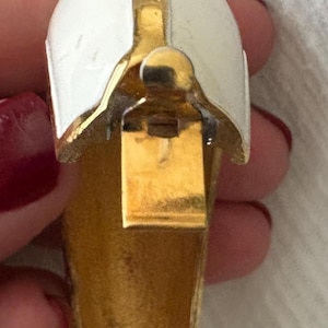 May include: A vintage gold-tone metal clip-on earring with a white enamel top. The earring has a curved, helmet-like design with a vertical gold stripe. The clip mechanism is visible.
