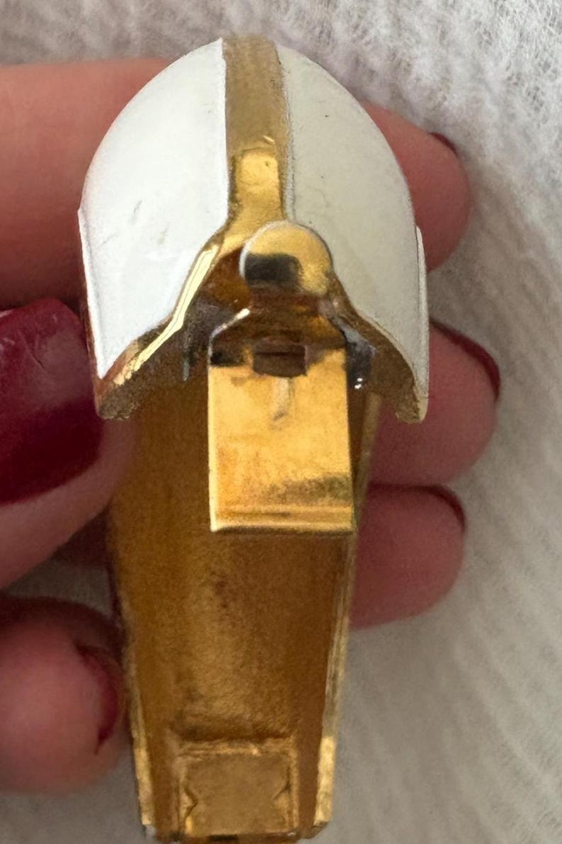 May include: A vintage gold-tone metal clip-on earring with a white enamel top. The earring has a curved, helmet-like design with a vertical gold stripe. The clip mechanism is visible.