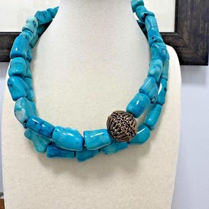 Può includere: Una collana di perline turchesi con una perlina decorativa color bronzo. La collana è esposta su un busto bianco. Le perline sono di forma irregolare e variano nelle tonalità di blu. La perlina di bronzo ha un motivo intricato.