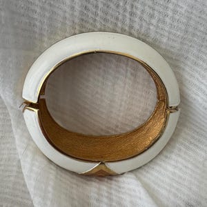 May include: A white and gold hinged bracelet. The bracelet has a rounded, oval shape with a white enamel exterior and a gold-toned interior. The clasp is gold-toned, and the bracelet is resting on a white fabric.