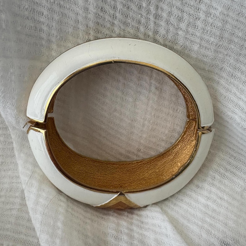 May include: A white and gold hinged bracelet. The bracelet has a rounded, oval shape with a white enamel exterior and a gold-toned interior. The clasp is gold-toned, and the bracelet is resting on a white fabric.