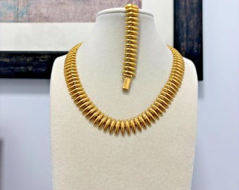 May include: A gold-colored necklace and bracelet set. The necklace and bracelet feature a repeating, ridged design. The jewelry is displayed on a white mannequin. The background includes a framed butterfly print.