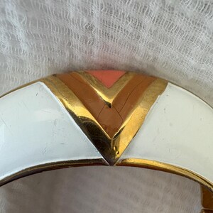 May include: A gold-toned hinged bracelet with a white enamel finish. The bracelet features a geometric design with a chevron pattern in shades of peach, brown, and gold. The bracelet is a vintage accessory.