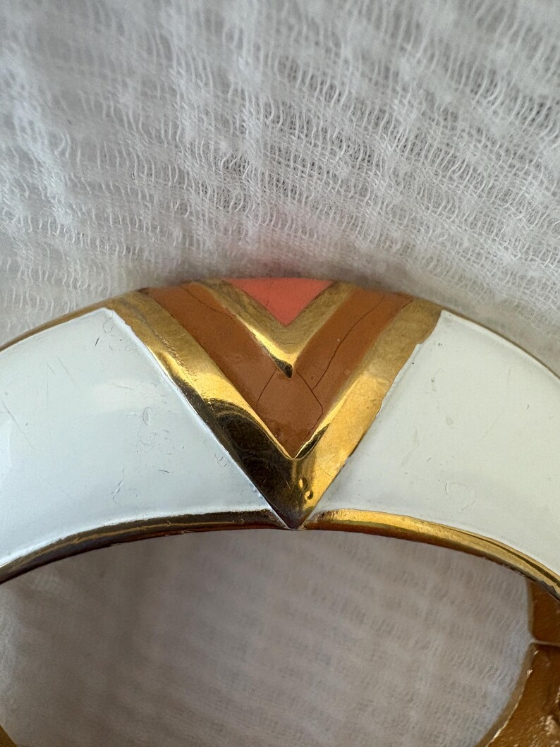 May include: A gold-toned hinged bracelet with a white enamel finish. The bracelet features a geometric design with a chevron pattern in shades of peach, brown, and gold. The bracelet is a vintage accessory.