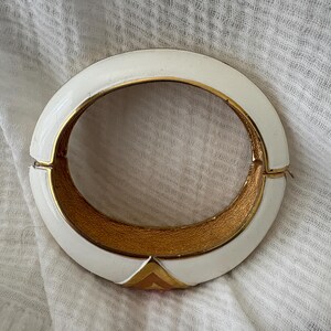 May include: White enamel hinged bracelet with gold-tone interior and accents. The bracelet features a geometric design with a gold triangle at the clasp. A vintage accessory.