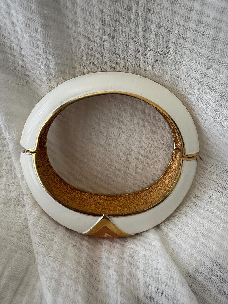 May include: White enamel hinged bracelet with gold-tone interior and accents. The bracelet features a geometric design with a gold triangle at the clasp. A vintage accessory.