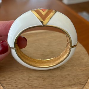 May include: White enamel hinged bangle bracelet with a gold interior and a geometric design in the center. The design features a gold chevron with a coral accent. A stylish piece of jewelry.