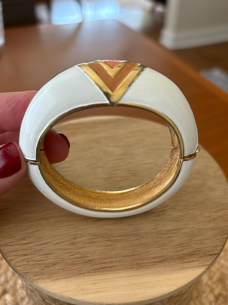 May include: White enamel hinged bangle bracelet with a gold interior and a geometric design in the center. The design features a gold chevron with a coral accent. A stylish piece of jewelry.