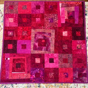 May include: A handmade quilt featuring a patchwork design in shades of red and pink. The quilt is composed of numerous squares and rectangles, with varying patterns and textures. The overall design is symmetrical, with a central focus. The quilt is bordered with a solid pink fabric.