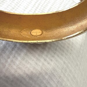 May include: A gold-toned bangle bracelet with a rounded shape. The interior of the bracelet has a textured finish and a small, oval-shaped stamp that reads "VALENTINO". The bracelet is set against a white, textured background.