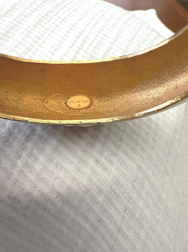 May include: A gold-toned bangle bracelet with a rounded shape. The interior of the bracelet has a textured finish and a small, oval-shaped stamp that reads "VALENTINO". The bracelet is set against a white, textured background.