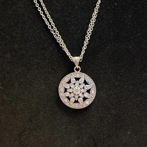 Pode incluir: Colar prateado com um pendente circular. O pendente apresenta um design floral com pedras preciosas claras e brilhantes. A corrente é delicada, sobre um fundo preto.