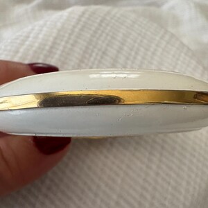 May include: A white compact case with a gold-colored band around the middle. The compact has a rounded shape and appears to be made of metal. The surface has a slightly textured appearance.