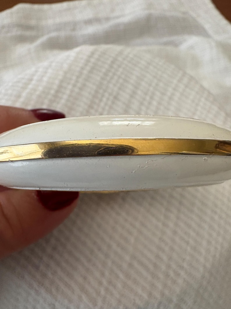 May include: A white compact case with a gold-colored band around the middle. The compact has a rounded shape and appears to be made of metal. The surface has a slightly textured appearance.