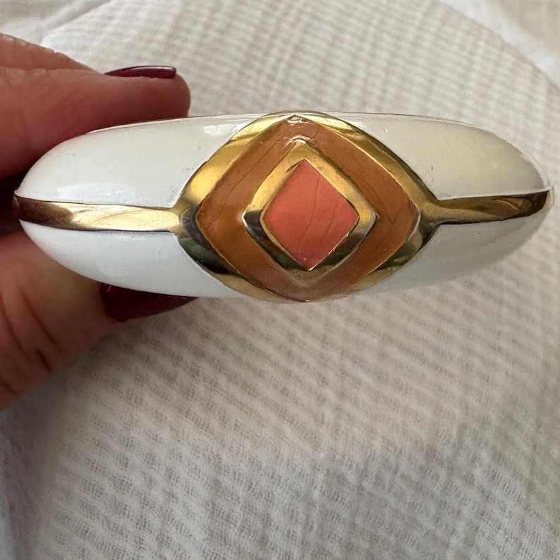 May include: White and gold-tone bracelet with a geometric design. The bracelet features a central coral-colored diamond shape, framed by a brown and gold border. The bracelet is oval-shaped and has a glossy finish.