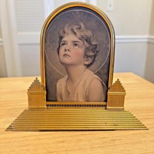 May include: A vintage gold-toned picture frame with a portrait of a child looking upwards. The frame has an arched top and a tiered base. The portrait is in black and white with a halo.