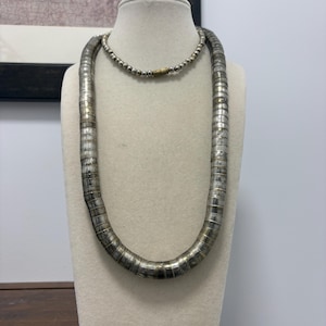May include: A long necklace featuring a thick, flexible chain composed of numerous flat, silver-toned metallic segments. The necklace includes a shorter, separate strand of small, round silver-coloured beads connected by a cylindrical brass-toned clasp.