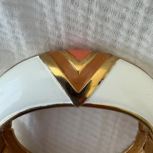 May include: A white and gold hinged bracelet with a geometric design. The bracelet features a white enamel exterior with gold accents and a central chevron pattern in gold, peach, and brown.