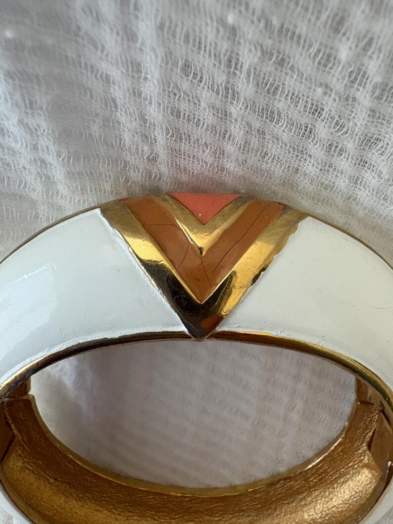 May include: A white and gold hinged bracelet with a geometric design. The bracelet features a white enamel exterior with gold accents and a central chevron pattern in gold, peach, and brown.