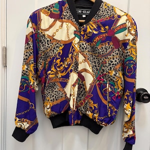 May include: A vintage bomber jacket with a striking pattern. The jacket combines purple, cream, gold, and brown hues with a leopard print and chain design. It has black cuffs, a black collar, and a zip closure. The label reads "Cline-Kolar".