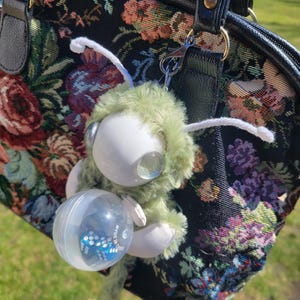 May include: A black floral tapestry handbag with a plush green and white toy attached. The toy has large, clear eyes and is holding a clear capsule containing blue dice. The handbag has black handles and trim.