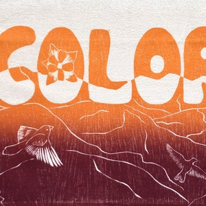 May include: A white and orange Colorado-themed textile features the word "COLORADO" in large, stylized letters. Below, a mountain range and flying birds are depicted in shades of orange and burgundy.