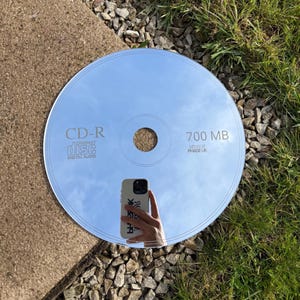 Peut inclure: Un disque CD-R réfléchissant avec le texte "CD-R compact disc digital audio" et "700 MB Mirror Phase UK". Le disque reflète le ciel et une main tenant un téléphone.