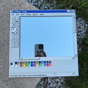 Può includere: Uno specchio quadrato progettato per assomigliare all'interfaccia del programma Windows Paint. La cornice grigio chiaro ha una barra del titolo blu e una tavolozza di colori nella parte inferiore. Il testo "untitled - Paint" è visibile in alto. Lo specchio riflette il cielo.