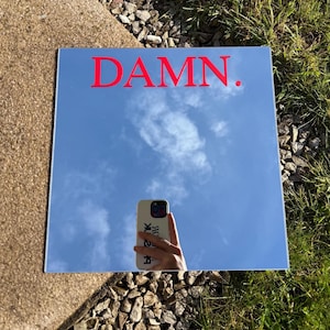 Può includere: Specchio quadrato con la scritta "DAMN." in lettere rosse. Lo specchio riflette un cielo blu con nuvole e una persona che tiene in mano un telefono. La custodia del telefono ha uno sfondo bianco con testo nero.