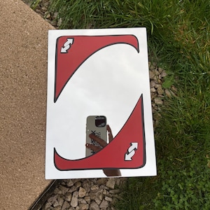 Può includere: Specchio rettangolare con sfondo bianco e grande design rosso. Il design rosso ha un contorno nero e presenta due frecce bianche che puntano in direzioni opposte. Lo specchio riflette l'ambiente.