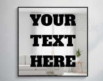 Op de afbeelding: Vierkante spiegel met zwart frame met de woorden "YOUR TEXT HERE" in vette zwarte letters. De spiegel reflecteert een witte bank, gordijnen en een kleine tafel met een plant.