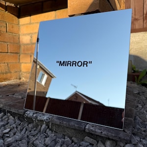 May include: A square mirror with the word "MIRROR" printed in black. The mirror reflects a clear blue sky and the surrounding environment, including a brick wall and a house.