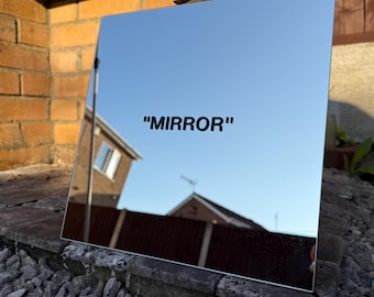 Op de afbeelding: Een vierkante spiegel met het woord "MIRROR" in het zwart gedrukt. De spiegel reflecteert een heldere blauwe lucht en de omgeving, inclusief een bakstenen muur en een huis.