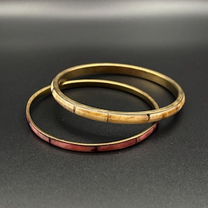 Puede incluir: Dos pulseras rígidas circulares con acabado metálico. Una pulsera presenta una incrustación lisa de color beige claro con líneas verticales oscuras, mientras que la otra muestra una incrustación veteada de color rosa rojizo segmentada.