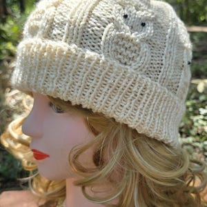 May include: A cream-colored knit beanie featuring a cabled texture and a prominent owl motif integrated into the pattern. The owl design includes small black beads for eyes. The hat has a ribbed cuff and is displayed on a mannequin head.