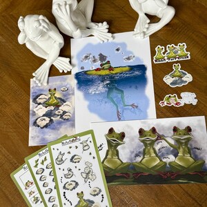 May include: An assortment of frog-themed art and decor. Includes white ceramic frog figurines, illustrated art prints, and sticker sheets. The prints and stickers feature cartoon frogs in various poses and settings, with text bubbles.