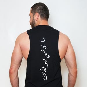 May include: Black sleeveless shirt with white Arabic script down the back. The shirt has a rounded hemline and a high neckline. The model is wearing olive green shorts and a watch with an orange band.