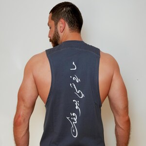 May include: Dark gray sleeveless shirt with white Arabic script on the back. The shirt has a scoop neck and wide armholes. The person is wearing a red and black watch.