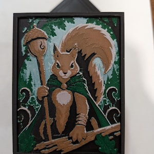 May include: A rectangular 3D printed wall plaque featuring a brown squirrel character wearing a green cape and holding a staff topped with an acorn. The design includes a dark background with green forest elements and white accents, framed in black.