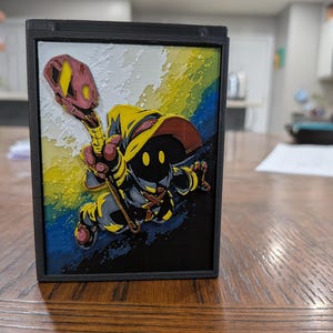 May include: A 3D-printed black frame featuring a colorful illustration of a character with a yellow hat and staff. The character is set against a background of rainbow colors, with a dark blue and yellow color scheme.