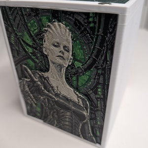 Könnte beinhalten: Eine weiße, rechteckige Aufbewahrungsbox mit einer 3D-gedruckten Reliefplatte auf der Vorderseite. Die Platte zeigt eine Figur mit texturiertem Haar und mechanischen Details vor einem grün-schwarzen Hintergrund mit Schaltkreismustern.