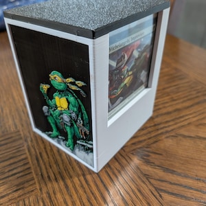May include: A white and black box with a textured grey top. The box features a Teenage Mutant Ninja Turtle graphic on one side and a card with text on another. The box is sitting on a wooden table.