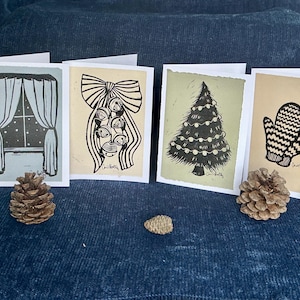 May include: Four handmade Christmas cards with winter-themed illustrations. Designs include a window with falling snow, a bow with bells, a decorated Christmas tree, and a pair of mittens. The cards are displayed on a blue surface with pine cones.