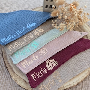 Puede incluir: Varios baberos personalizados para bebés, en colores como azul, beige y rosa. Cada babero tiene un nombre y pequeños dibujos, como un coche o un arcoíris. Los baberos son de tela texturizada y están colocados sobre una rodaja de madera.