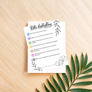 May include: A white card with the text "Bible Highlighting Color Code" and a list of colour-coded categories for Bible study. The card includes floral illustrations and is set on a wooden surface with a green leaf.