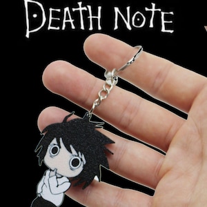 May include: A Death Note keychain featuring a cartoon character with black hair, a white shirt, and black pants. The keychain is held against a black background, with the words "Death Note" in white above.
