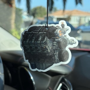 May include: An air freshener shaped like a detailed illustration of a car engine. The engine is primarily gray with black accents and a touch of orange. It hangs from a black string, designed for use in a vehicle.