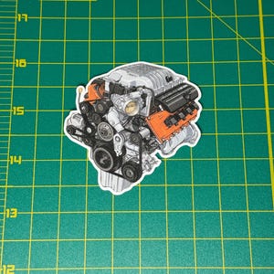 May include: A sticker featuring a detailed illustration of a car engine, with gray, black, and orange components. The engine is a complex design, showcasing belts, pulleys, and various mechanical parts. The sticker is approximately 4 inches in length.