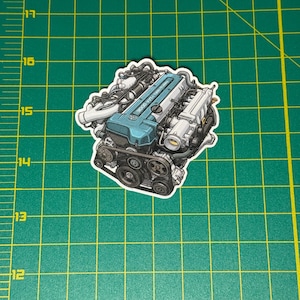 May include: A sticker featuring a detailed illustration of a car engine. The engine has a blue valve cover with the text "3000 VVT-i Twin Turbo". The sticker is on a green cutting mat with inch markings.
